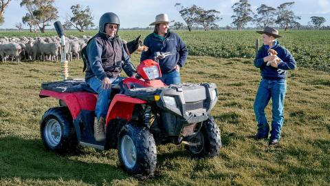 A farmer wearing a helmet on a quad bike, talking to two people standing nearby